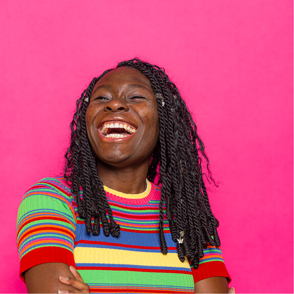 Smiling woman with braided hair wearing a colorful striped shirt against a bright pink background.