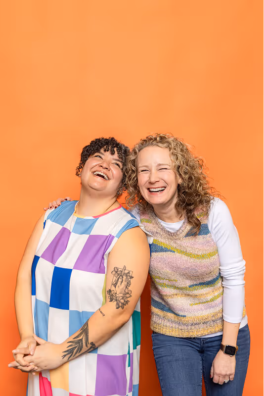 Two women smiling and leaning close against an orange background, one with curly dark hair and colorful checkered dress showing tattoos on her arm, the other with wavy blonde hair wearing a striped knitted vest over a white long-sleeve shirt.
