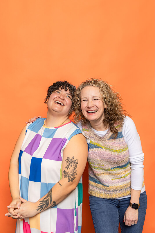 Two women smiling and leaning close against an orange background, one with curly dark hair and colorful checkered dress showing tattoos on her arm, the other with wavy blonde hair wearing a striped knitted vest over a white long-sleeve shirt.