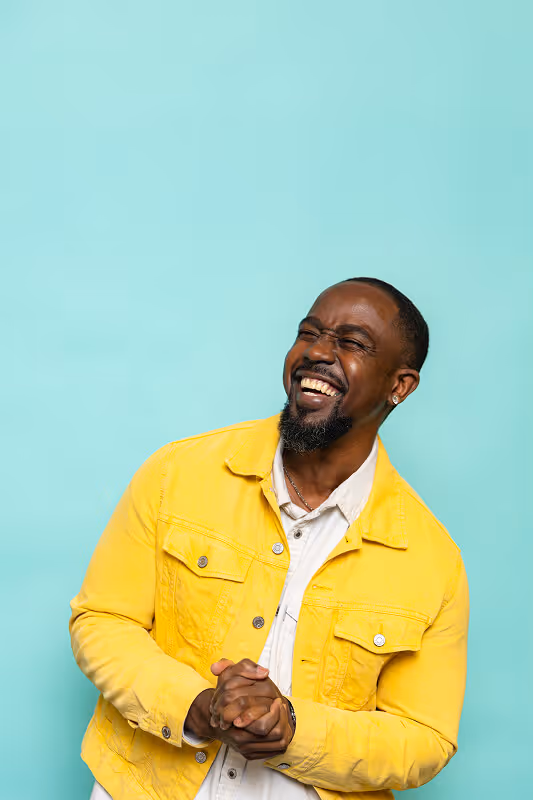 Smiling man in a yellow jacket clasping his hands against a light blue background.