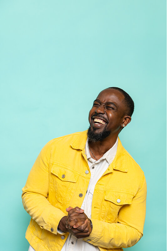 Smiling man in a yellow jacket clasping his hands against a light blue background.