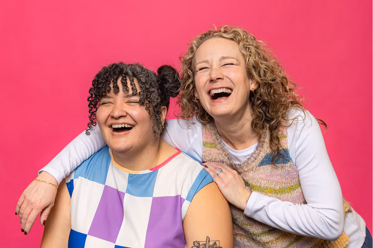 Two women laughing together in front of a bright pink background, one with curly hair wearing a colorful checkered top and the other with blonde curly hair in a knitted vest.