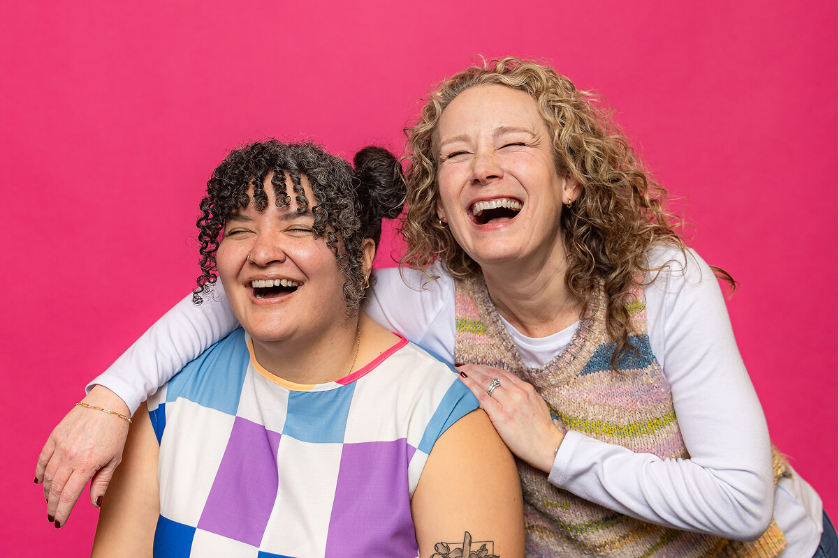 Two women laughing together in front of a bright pink background, one with curly hair wearing a colorful checkered top and the other with blonde curly hair in a knitted vest.