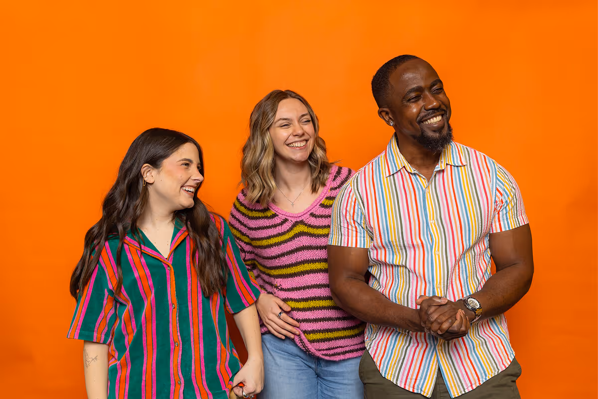 Three smiling people wearing colorful striped and patterned tops standing against an orange background.