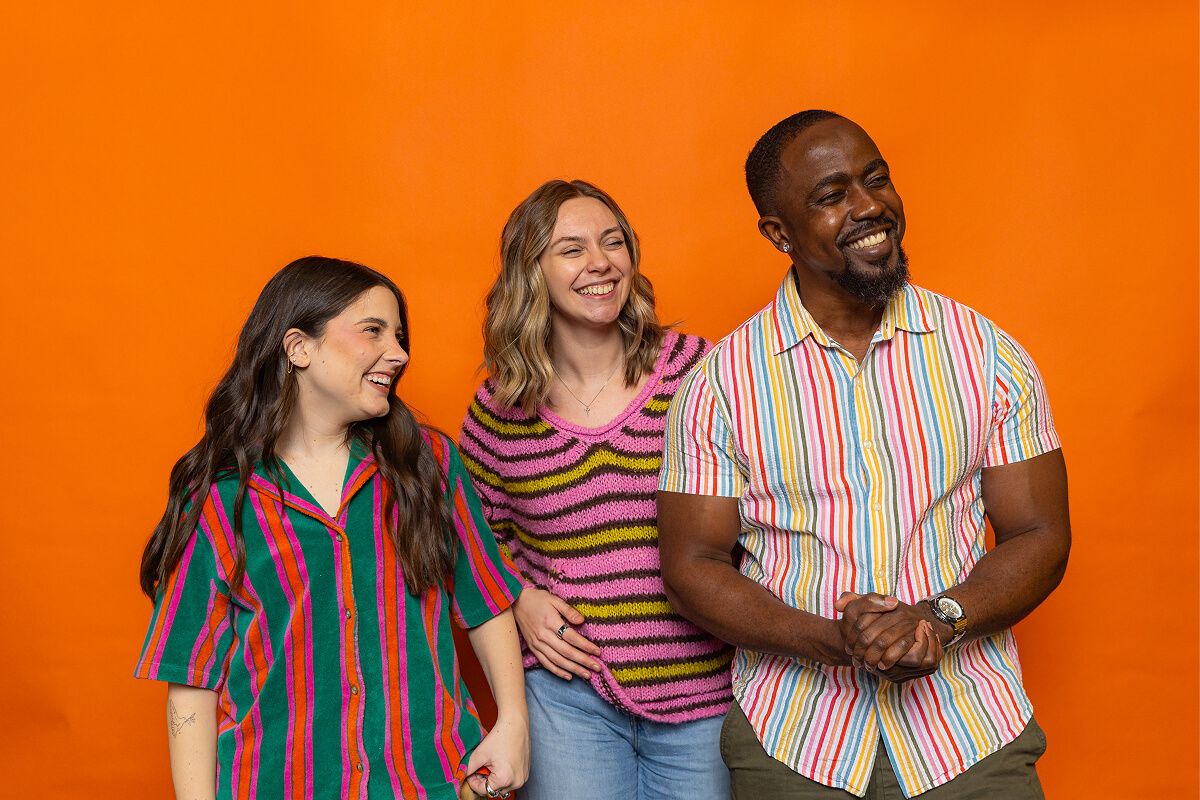 Three smiling people wearing colorful striped and patterned tops standing against an orange background.