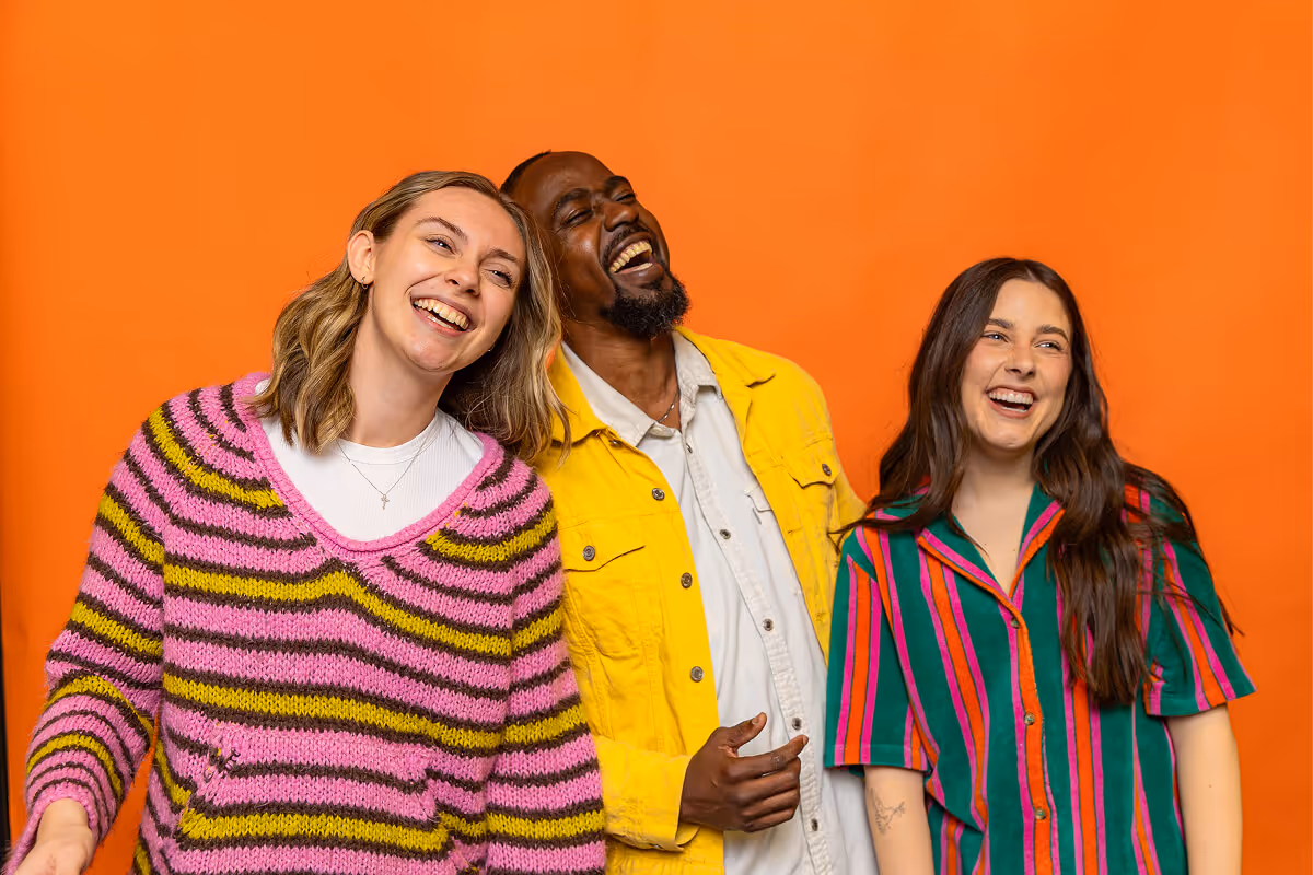 Three diverse friends laughing and standing closely together against an orange background.