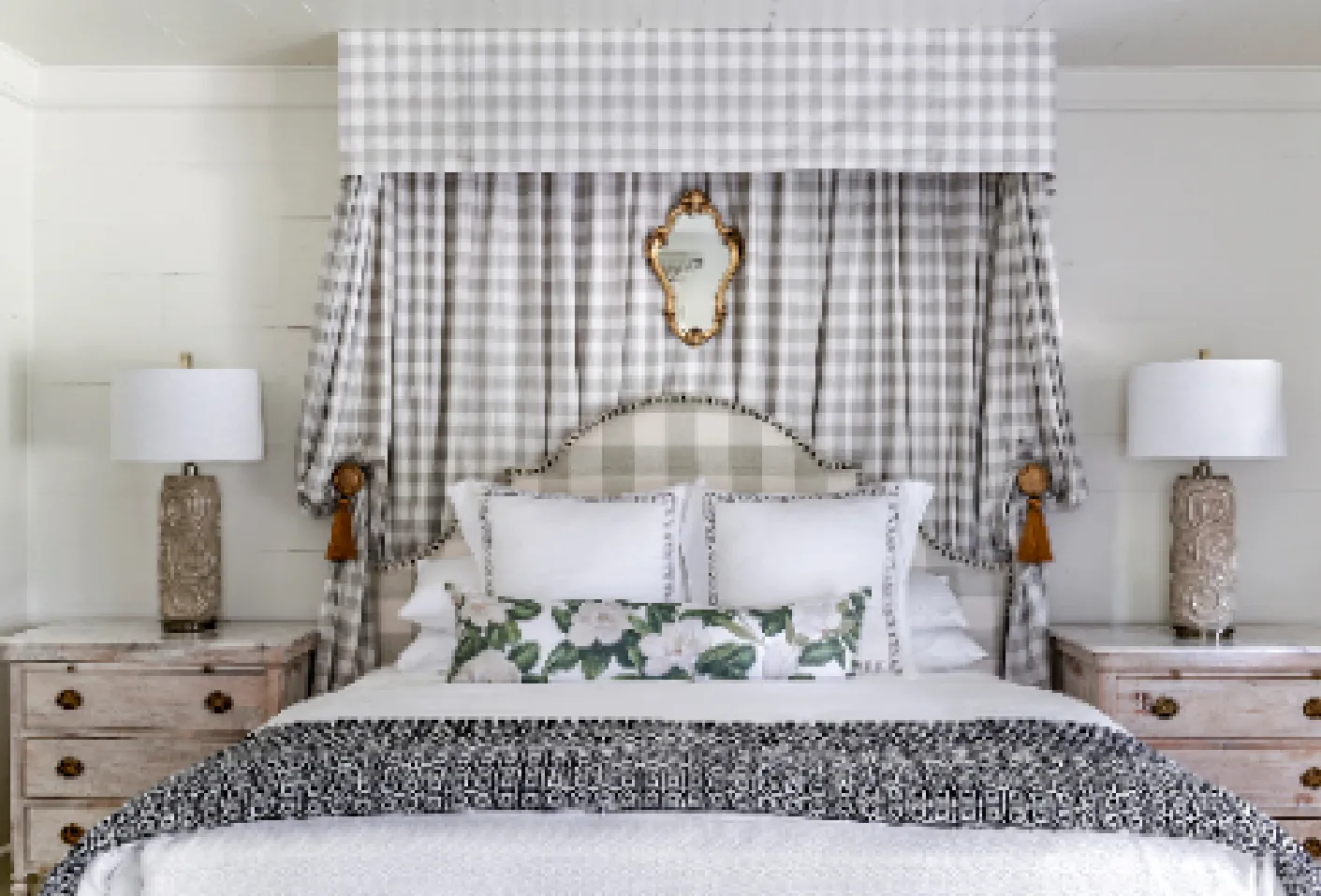 Bedroom with a checkered fabric canopy, checkered headboard, white pillows with trim, floral lumbar pillow, patterned throw blanket, two rustic nightstands, and ornate table lamps.