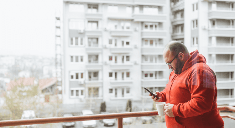 Man on balcony looking at his phone and holding a cup of coffee