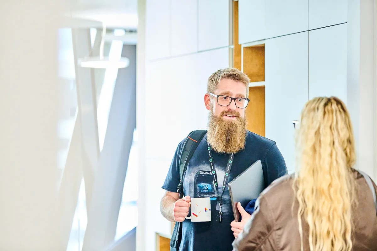Two Leighton colleagues stood in a hallway chatting. One is holding a laptop and a coffee mug, the other has her back to the camera.