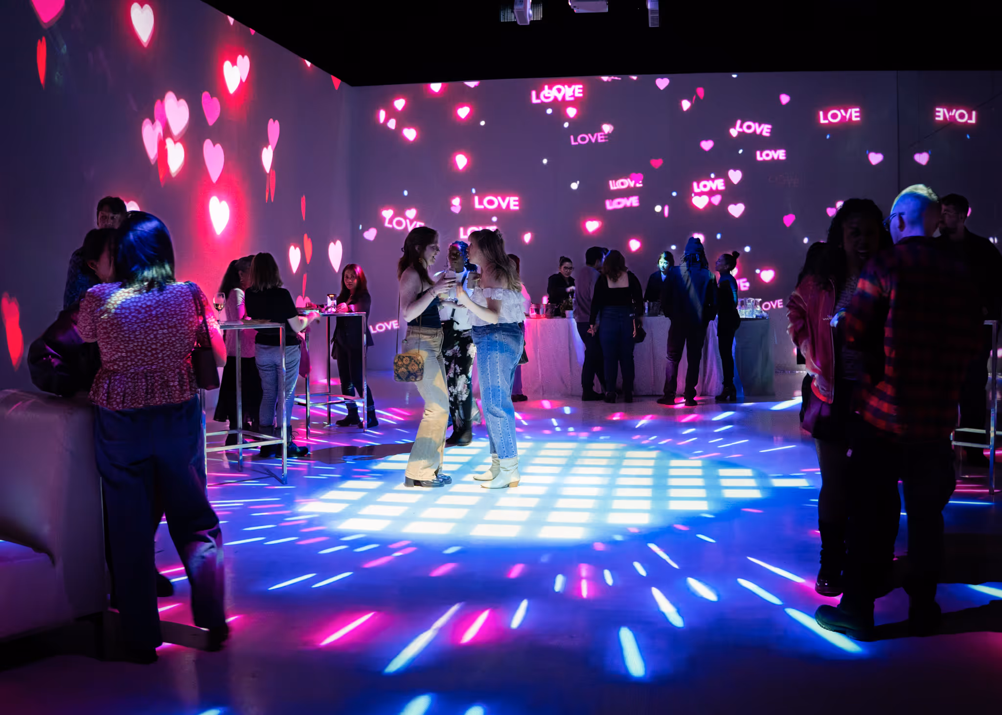 People socialize at NATM event with pink heart-shaped and "LOVE" light projections on the walls and blue spotlights on the floor.