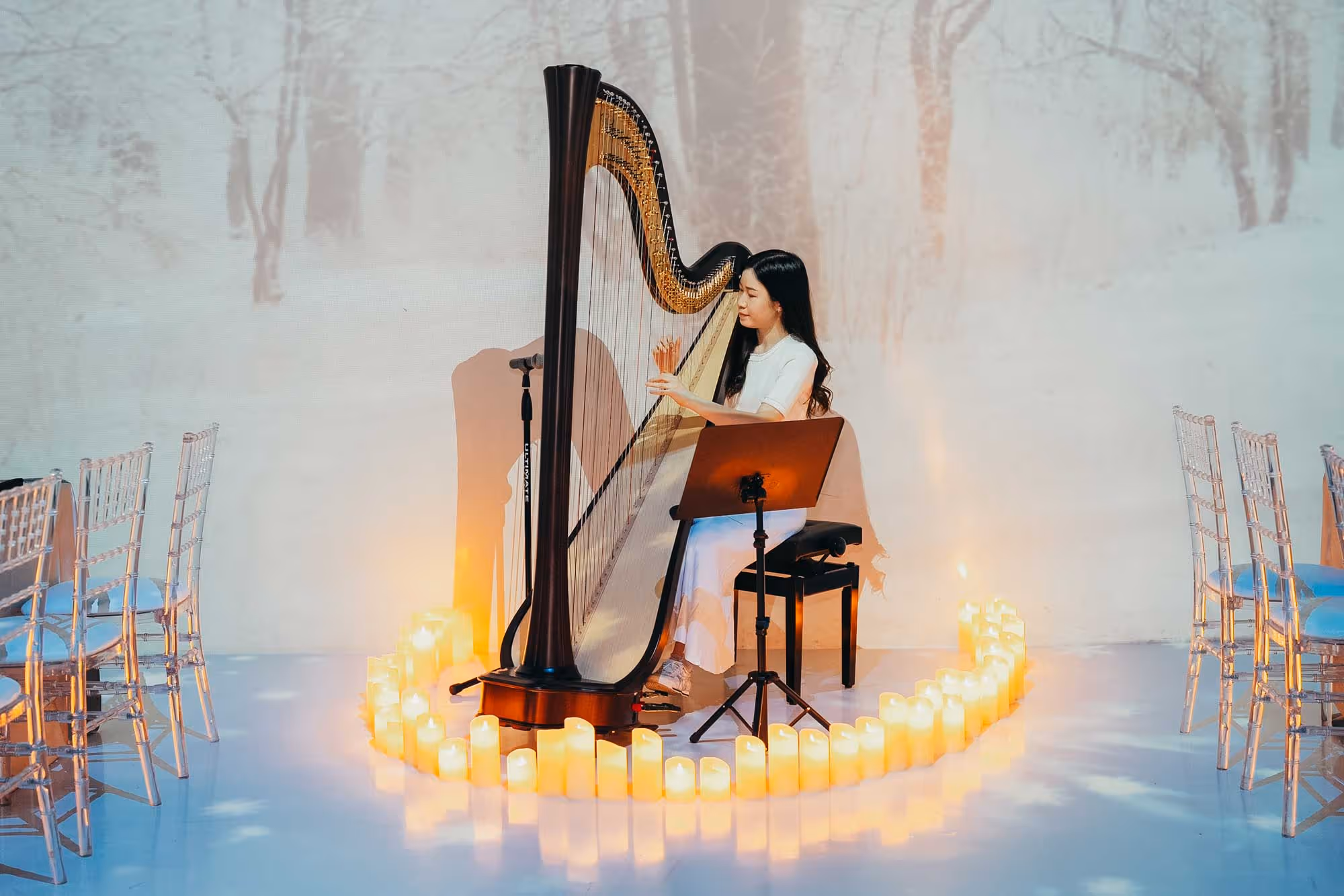 A woman plays a harp surrounded by glowing candles on a white floor, with a snowy forest backdrop. The scene is serene and elegant, evoking calm.
