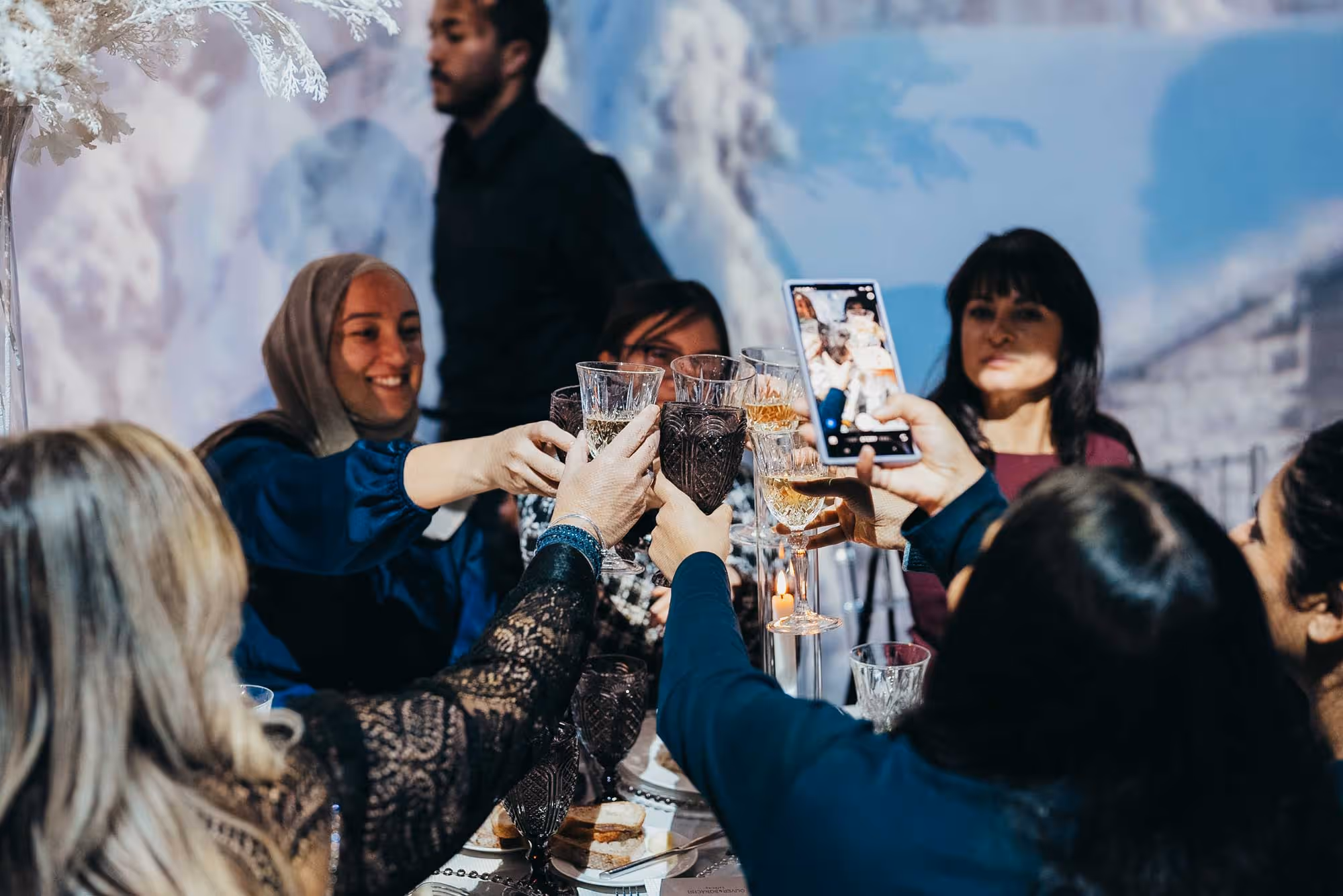 A group of people toasting with glasses around a table, smiling and capturing the moment with a phone. The atmosphere is festive and joyful.