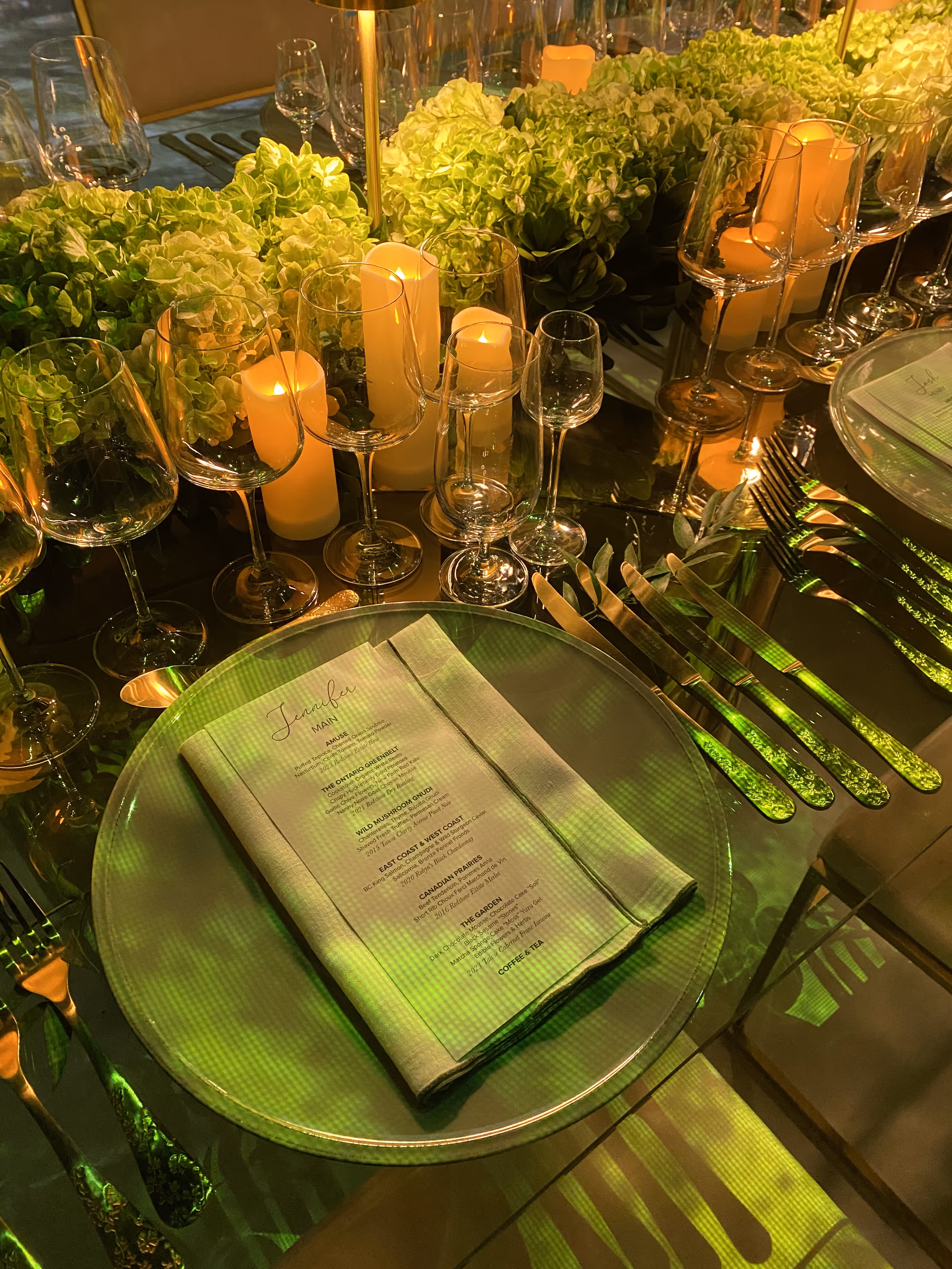 Elegant table setting with lush green hydrangeas, lit candles, and glassware. A menu rests on a reflective plate, conveying a warm, sophisticated ambiance.