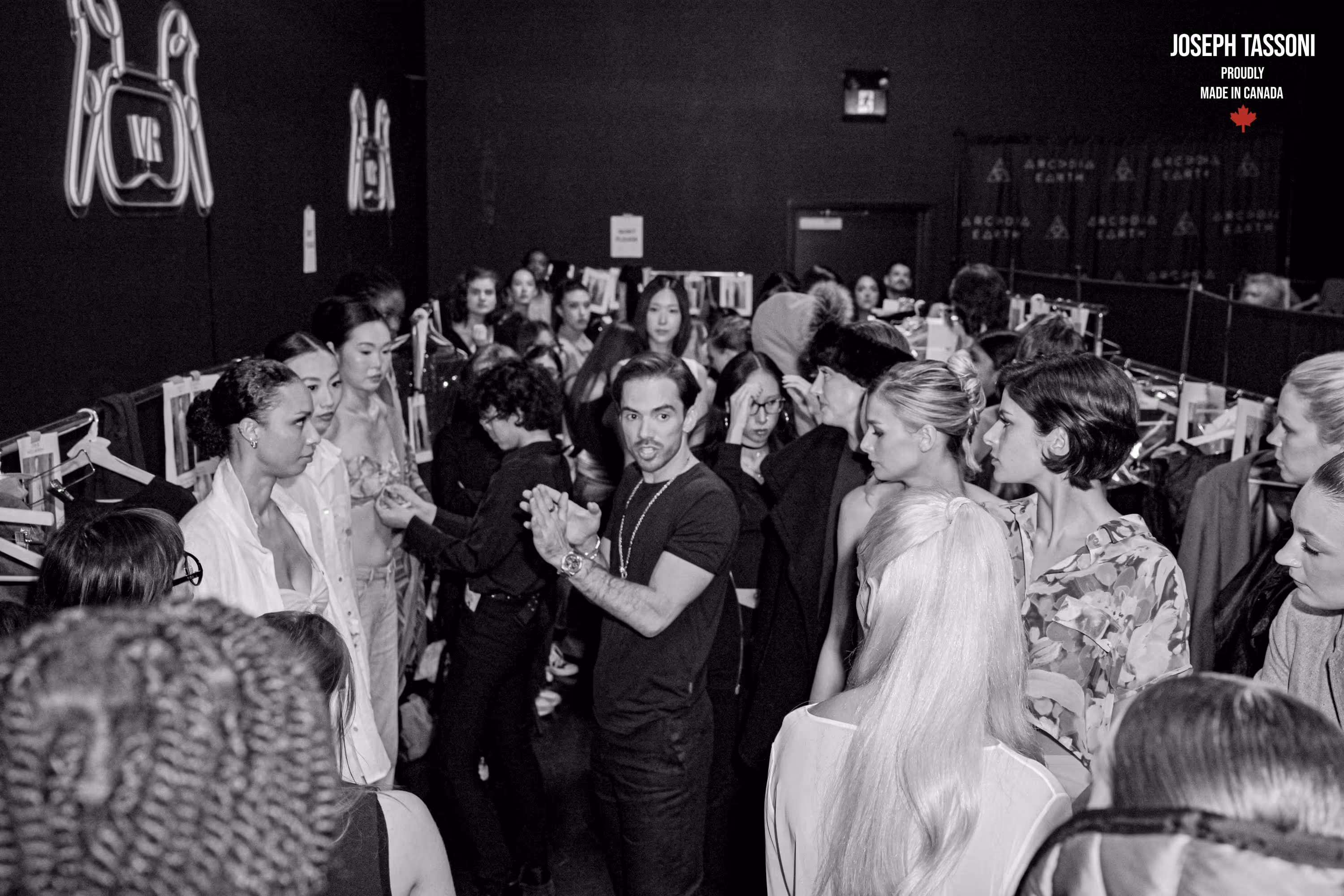 Black and white image of a busy backstage scene at a fashion show. Models gather around a director, receiving instructions. The atmosphere is focused and energetic.