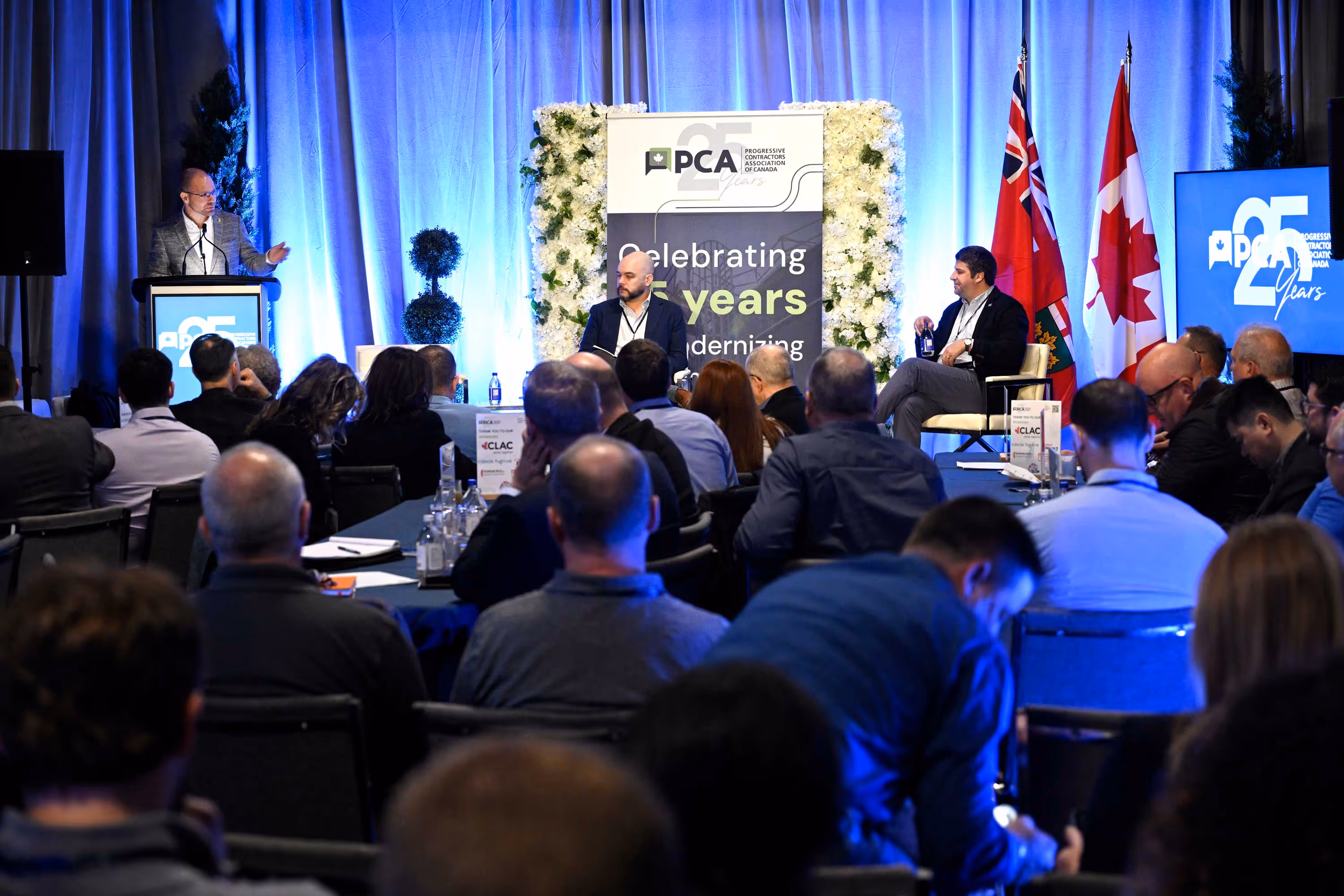 A conference room with a speaker at a podium on the left and two men seated on stage discussing. Attendees face forward. Canadian flags are visible, with a backdrop displaying "PCA 25 Years." The atmosphere is formal and engaged.