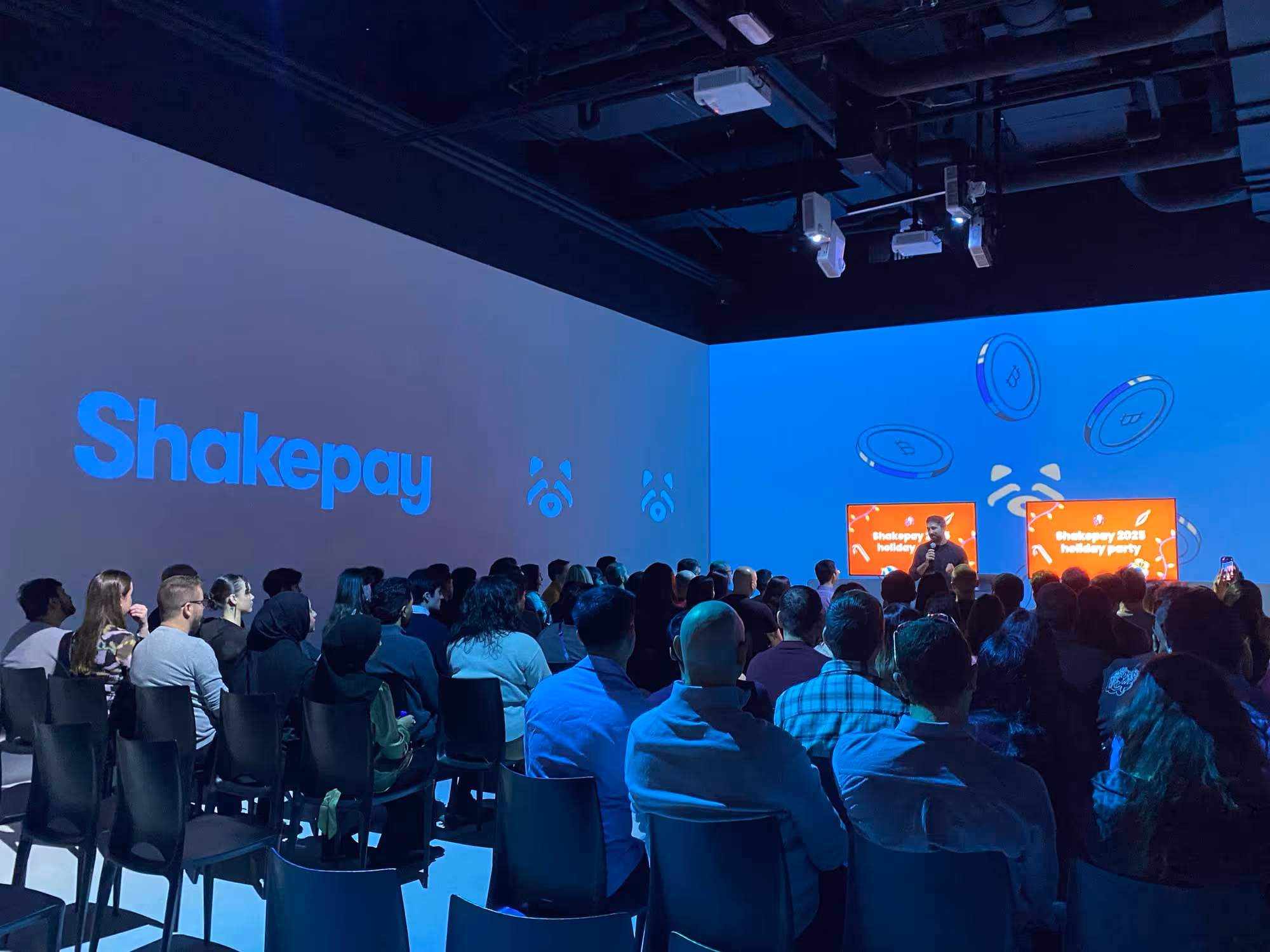 Audience in a dimly lit room faces a speaker on stage with screens displaying "Shakepay 2025 Holiday Party." Blue lighting sets a tech-focused ambiance.