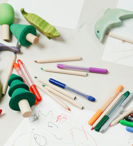 Colorful markers, pens, and wooden toys shaped like trees, a pea pod, and a dolphin scattered around a child's scribbled drawing on white paper.