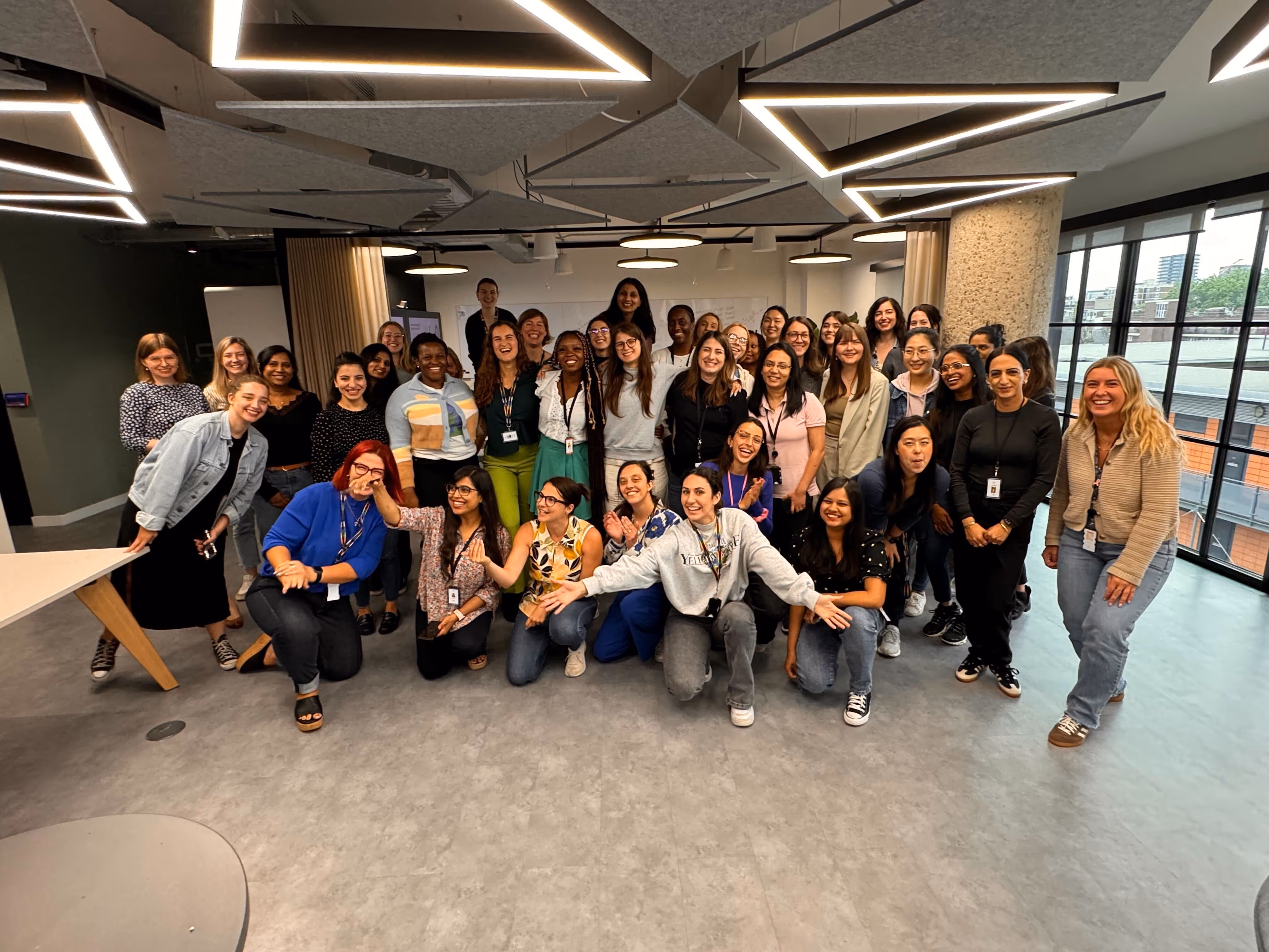 Group of diverse women smiling and posing inside a modern office space with large windows and geometric ceiling lights.