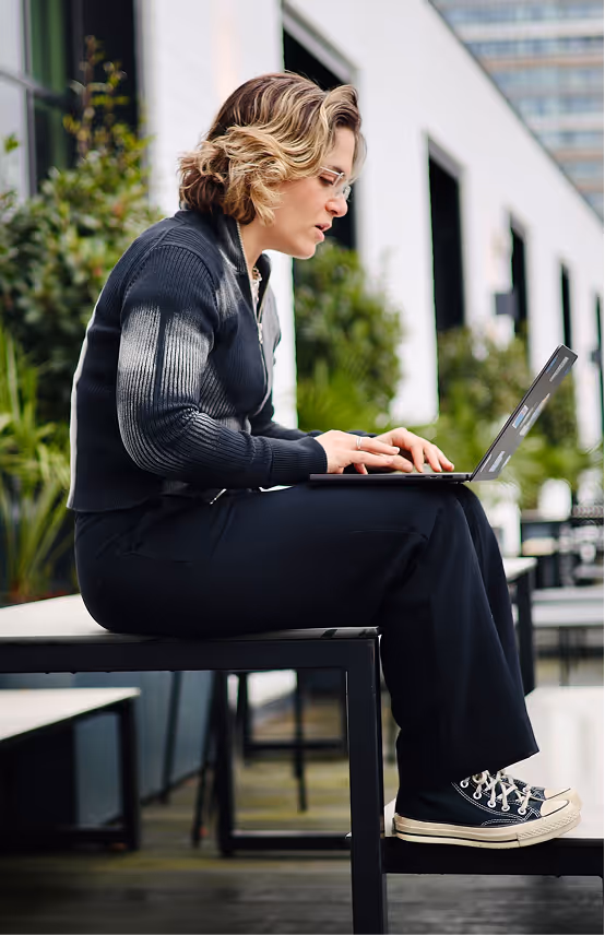 Woman with short wavy hair and glasses sitting on a bench outside, working on a laptop.