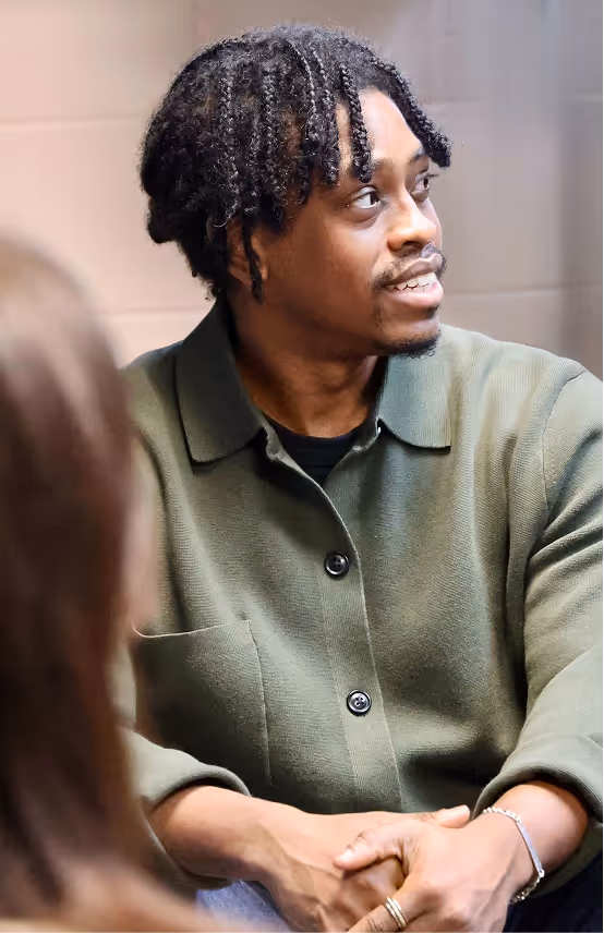 Man with braided hair wearing a green shirt looking to the side while speaking with another person.
