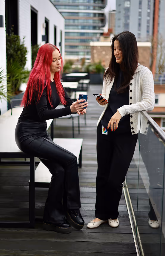Two young women outdoors on a balcony, one with long red hair sitting and showing her phone, the other with long black hair standing and holding a phone.