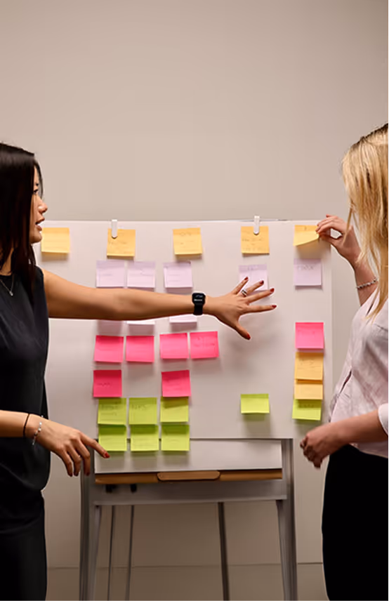 Two women interacting and discussing colorful sticky notes arranged on a whiteboard.