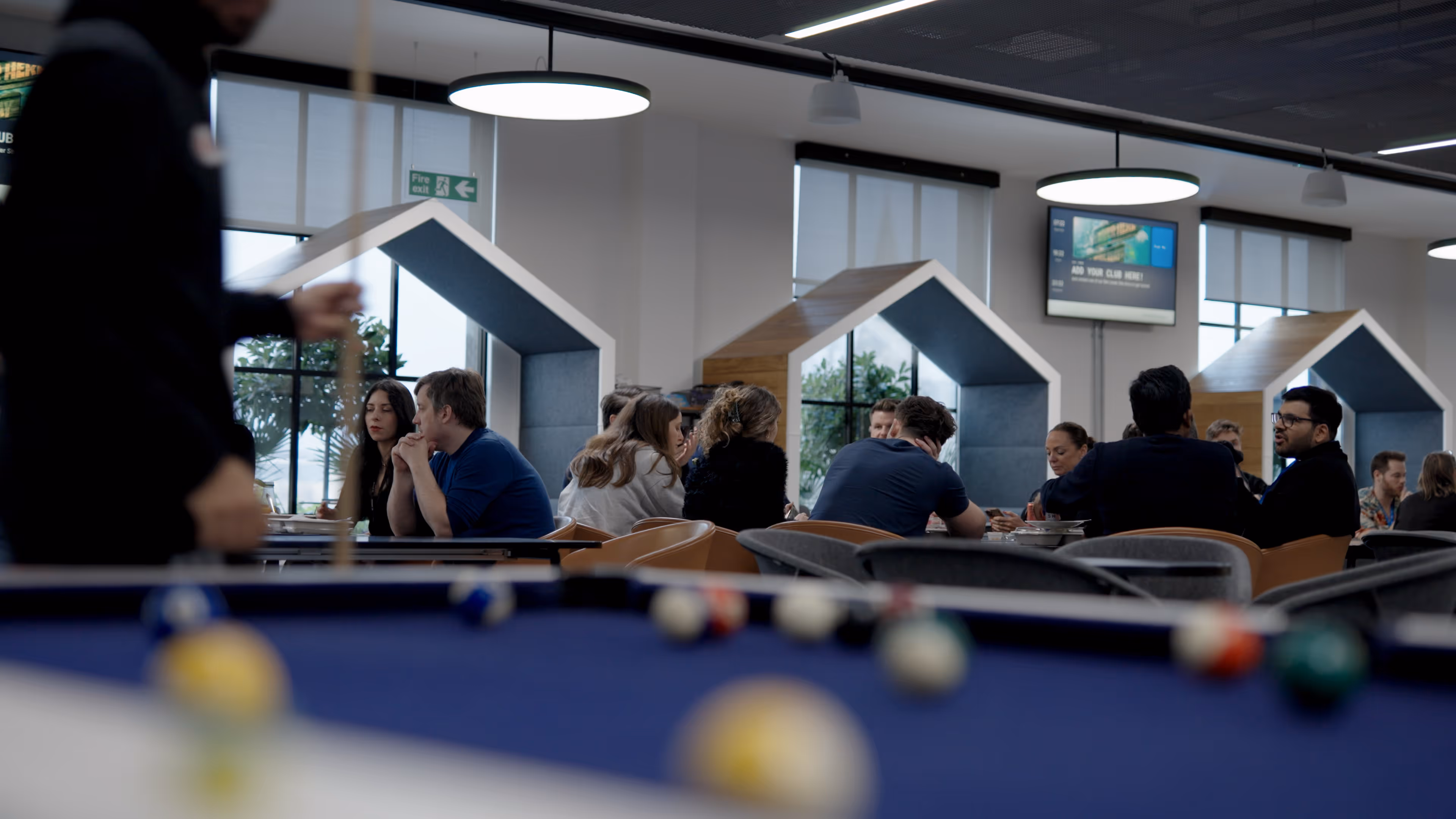 Group of people sitting and talking at tables with house-shaped booths in a modern indoor space, with a person playing pool in the foreground.