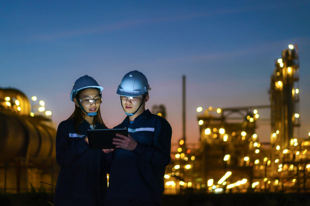 Zwei Fachkräfte mit Schutzhelmen und reflektierender Arbeitskleidung stehen vor einer beleuchteten Industrieanlage bei Dämmerung und betrachten gemeinsam ein Tablet.“