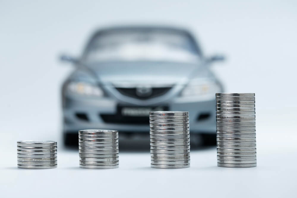 Verschwommenes silbernes Auto im Hintergrund, im Vordergrund vier Münzstapel mit zunehmender Höhe von links nach rechts.
