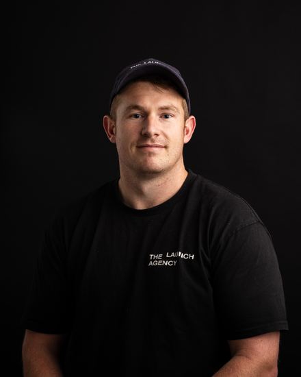 Portrait of Ben Langton Brunell, CEO & founder of The Launch Agency, wearing a black cap and a black t-shirt with the text 'THE LAUNCH AGENCY' on it, against a black background.