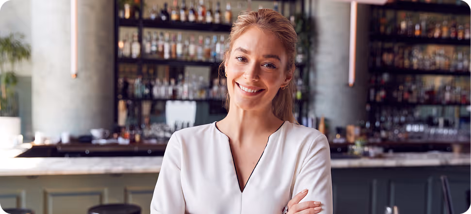 Smiling woman with folded arms standing in a bar with shelves of bottles in the background.
