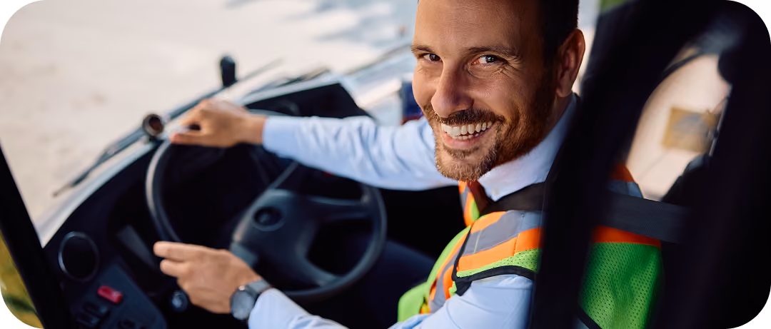Smiling man wearing a green safety vest and white shirt sitting in the driver's seat of a vehicle, holding the steering wheel and looking at the camera.