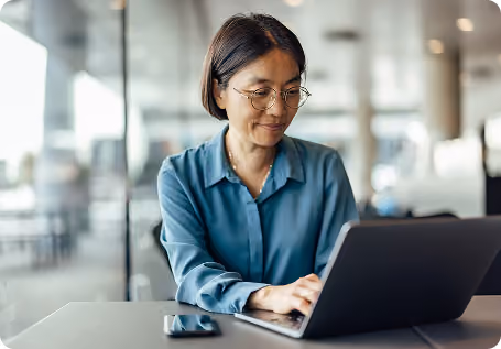 Person wearing glasses and a blue shirt typing on a laptop at a desk with a smartphone nearby, in a modern office setting.