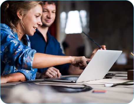 Two people sitting at a table working together on a laptop, with the woman pointing at the screen and the man watching.