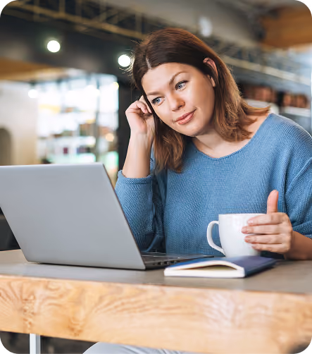 A woman with shoulder-length brown hair wearing a blue sweater looks thoughtfully at a laptop screen while holding a white mug in one hand, sitting at a wooden table with a closed notebook in front of her in a well-lit indoor space.