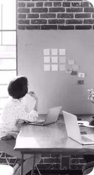 Two people sitting at a table with laptops, one person looking thoughtfully at sticky notes arranged on a wall in a work environment with brick wall and window.