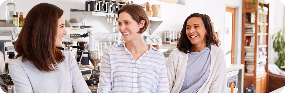 Three women smiling and talking together in a modern kitchen with shelves and kitchenware.