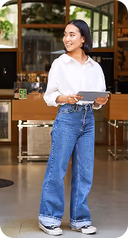 Young woman with short black hair wearing a white blouse and blue jeans, standing inside a modern cafe holding a tablet and pen, smiling while looking to the side.