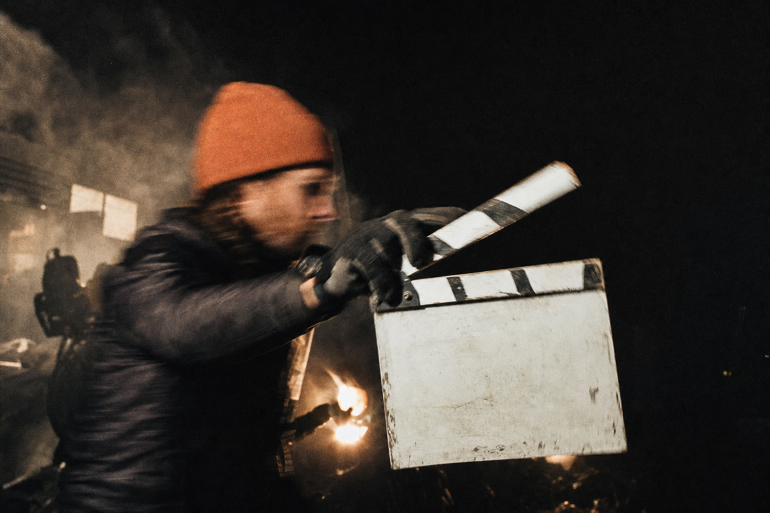 A crew member in a rust-orange beanie snaps a blank film clapperboard shut on a hazy night shoot, arm blurred with motion, lights glowing behind.
