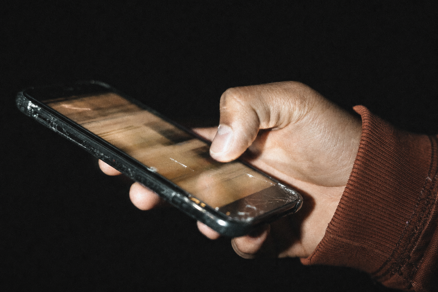 Hard flash catches a hand scrolling a phone in a pitch-dark room, thumb mid-swipe over a cracked screen that is only a smear of warm light.