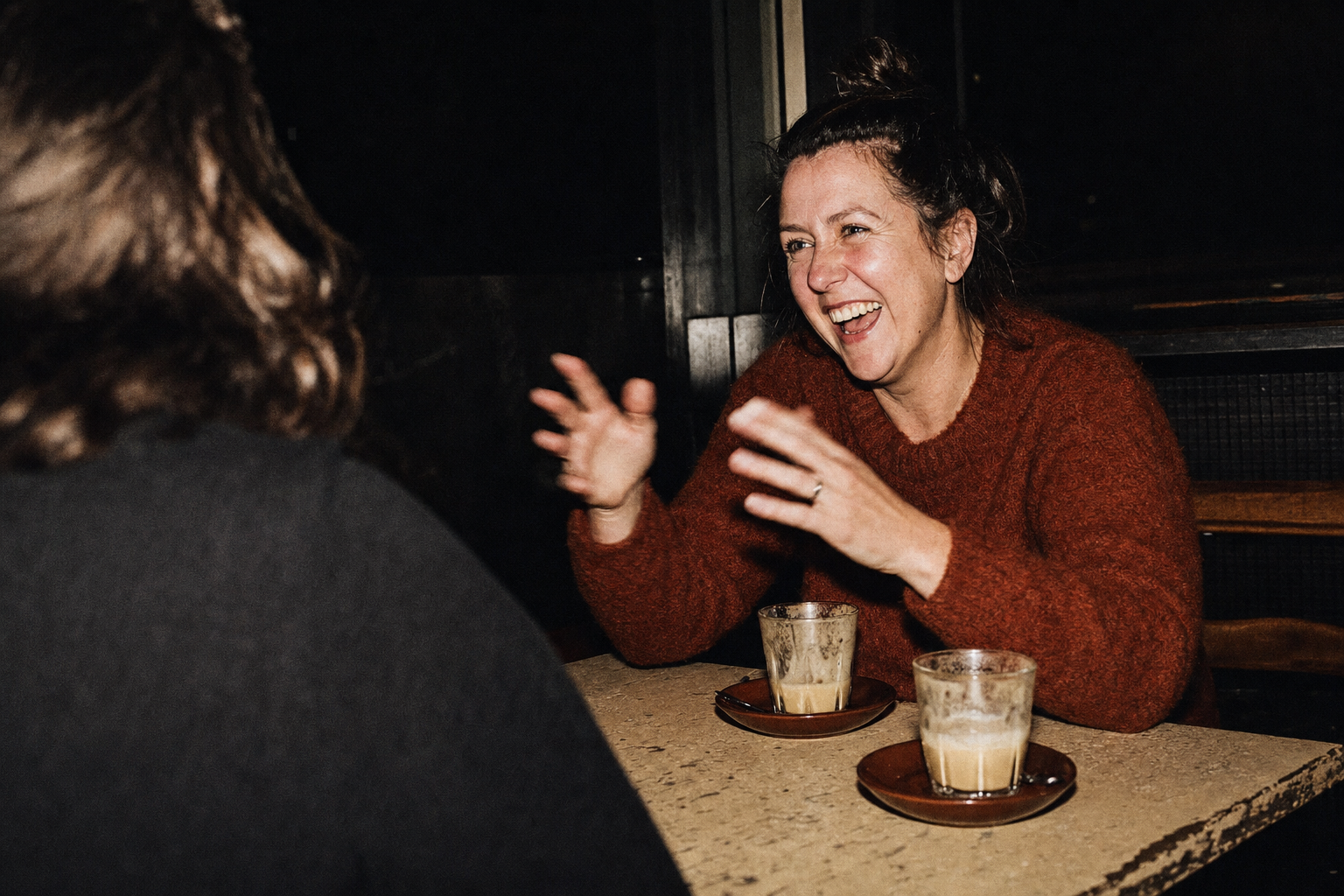A woman in a rust-red jumper laughs mid-story across a chipped cafe table at night, hands blurred with gesture, two half-drunk flat whites lit by hard flash.