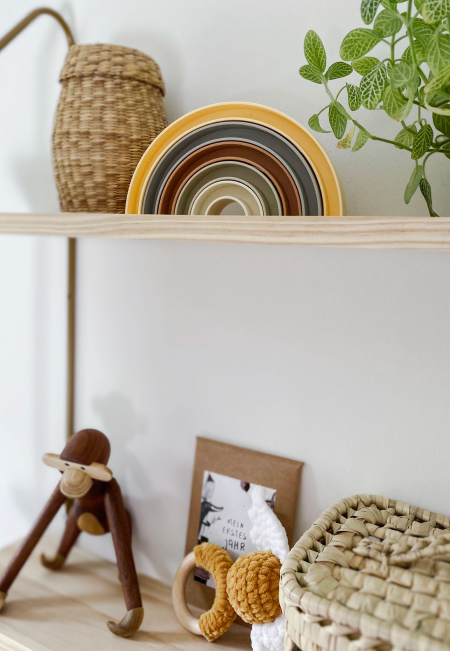 Ein Holzregal an der Wand mit unterschiedlichen Deko-Artikeln – wie z.B. einem Affen aus Holz, ein Regenbogen zum Stecken oder kleinen Körbchen.