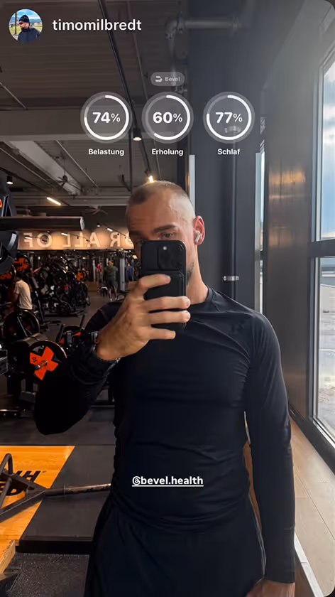 Man in black workout clothes taking a mirror selfie in a gym, with fitness stats showing 74% Belastung, 60% Erholung, and 77% Schlaf at the top.