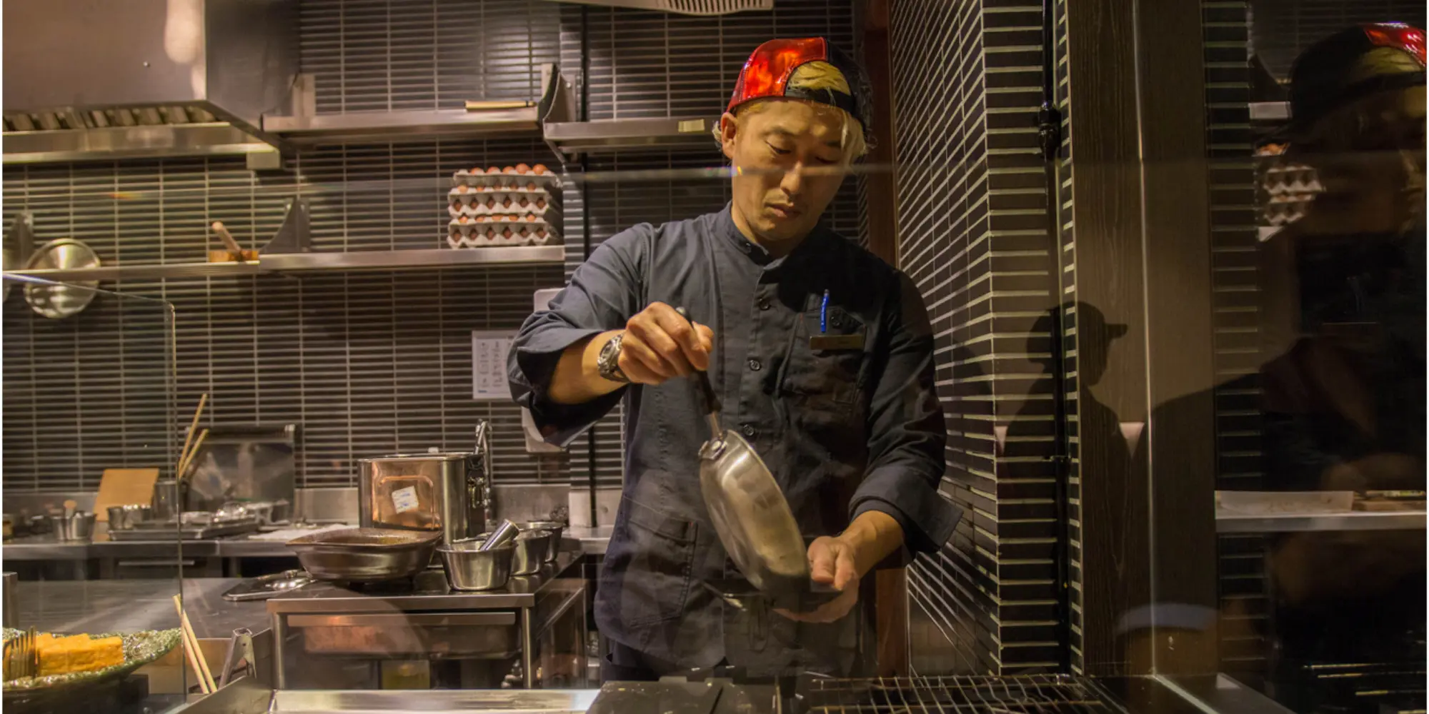 Chef wearing a red cap and dark jacket preparing food in a professional kitchen with cooking utensils and egg trays in the background.