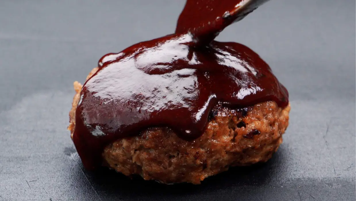 Grilled burger patty being topped with thick dark barbecue sauce on a flat surface.