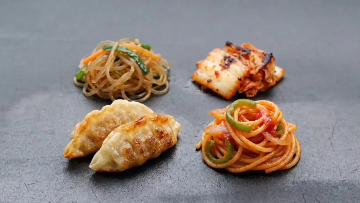 Plate with two fried dumplings, a serving of glass noodles with vegetables, kimchi, and seasoned spaghetti with green bell peppers.