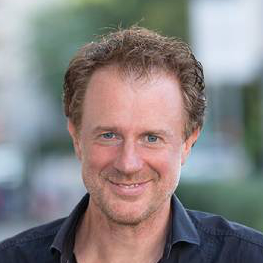 Manfred Gansterer, Agentur Grundauf, Smiling man with short curly brown hair and blue eyes wearing a dark shirt, outdoors with blurred background.