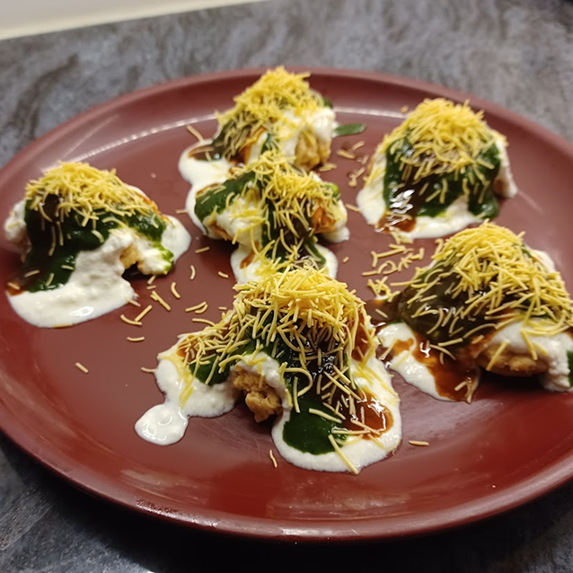 Plate with six pieces of Indian snack dahi puri topped with white yogurt, green chutney, tamarind chutney, and sev.