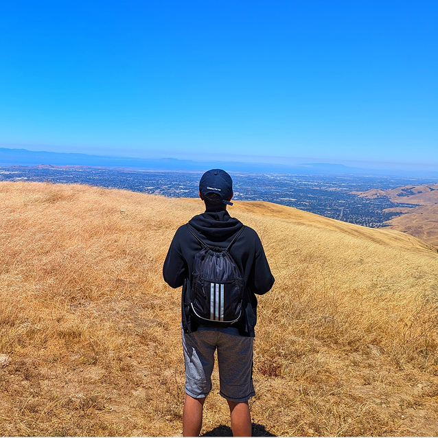 Naman wearing a black hoodie and backpack standing on a dry grassy hill overlooking a cityscape under a clear blue sky.