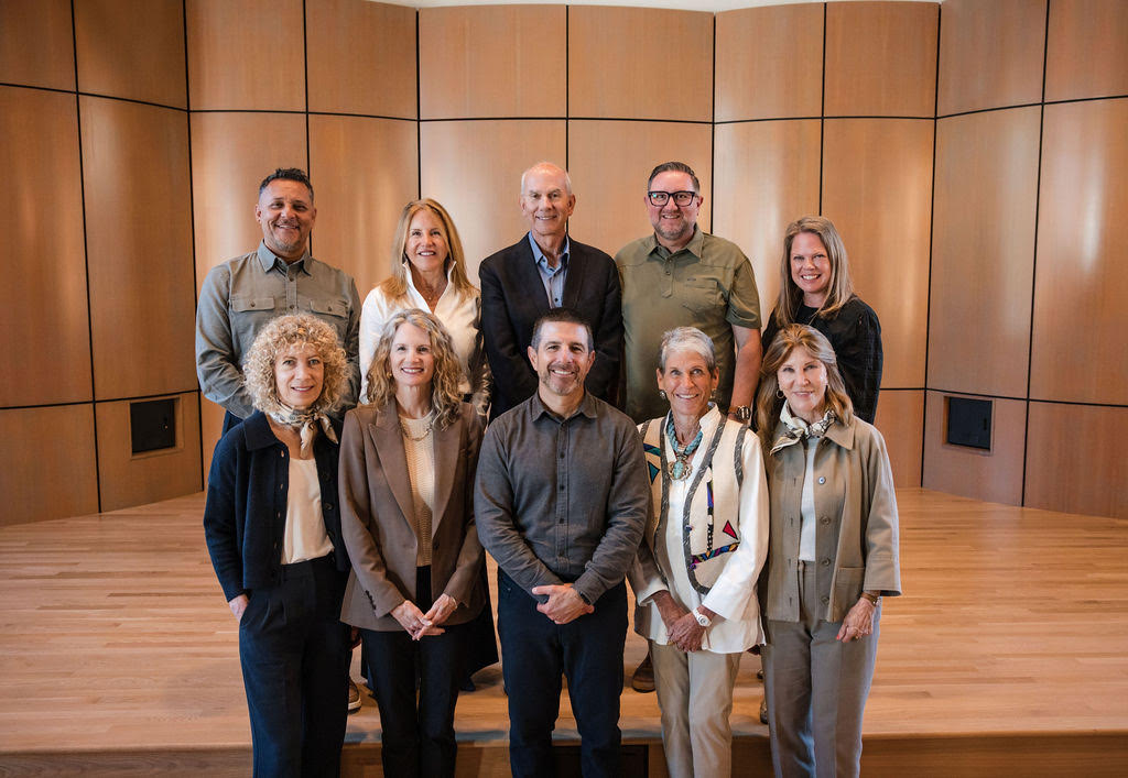 Group of ten professionally dressed adults standing on a wooden stage in front of a wood-paneled wall, smiling at the camera.